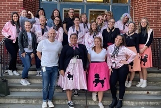 Teachers and staff standing on school stairs wearing 50s themed halloween costumes such as pink poodle skirts, black leather jackets and pink lady jackets