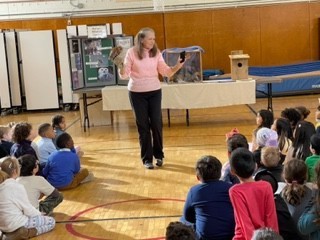 woman in pink shirt with a live owl on her arm presenting to a group of elementary students in a school gymnasium