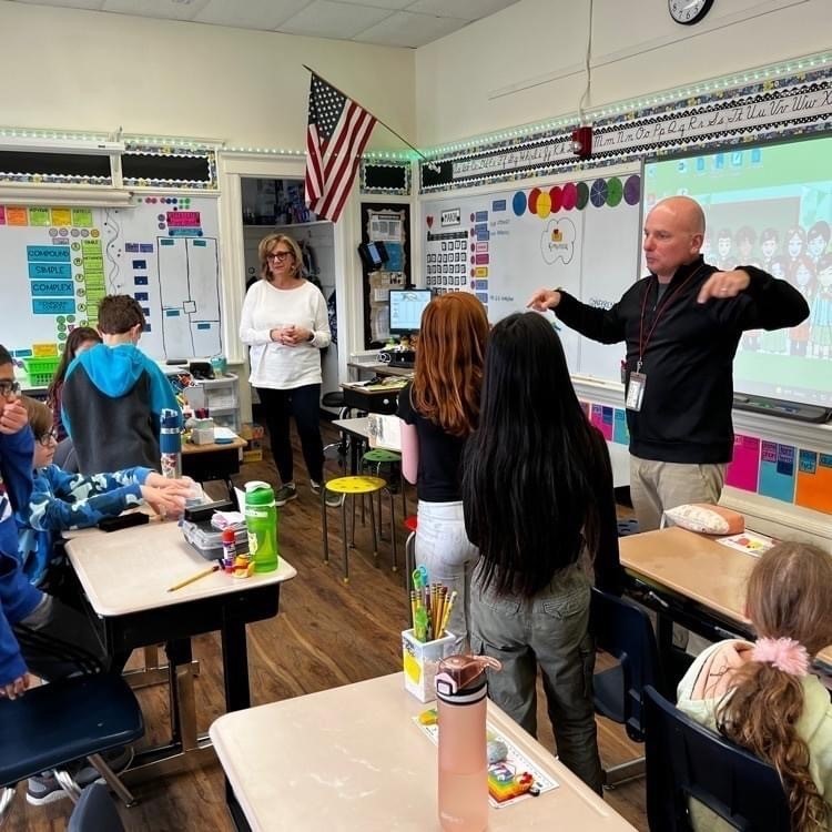 bald man in a black shirt standing in front of a smartboard with a group of 4th graders and their teacher in their classroom