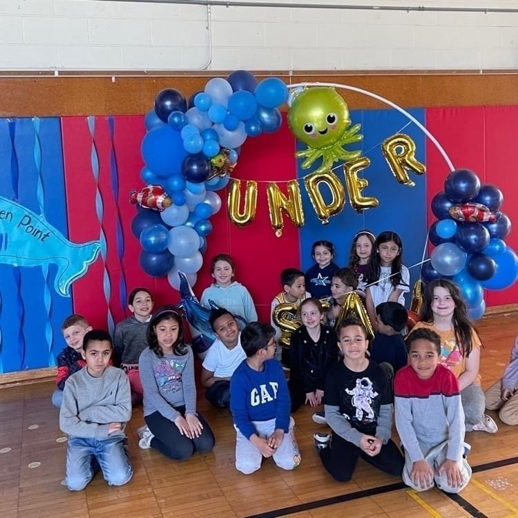 about 12 students sitting on a floor in from of a ballon arch that says under the sea