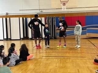 two basketball players dressed in black and two students stand in front of classmates  in the school gymnasium