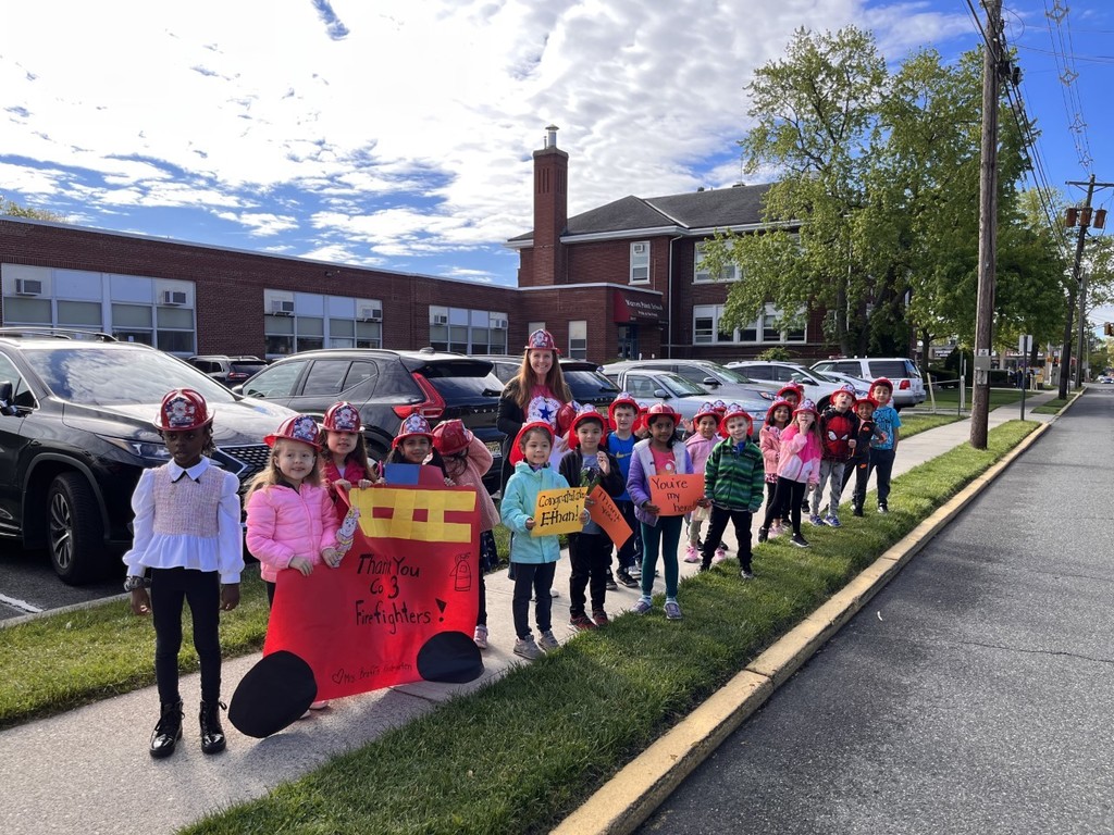 Kindergarten students stand outside of school with red fire hats on holding posters