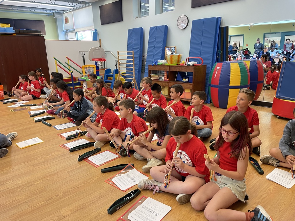 Third grade students and teacher wearing red shirts playing recorders and reading music as they sit on the floor in a school gymnasium. 