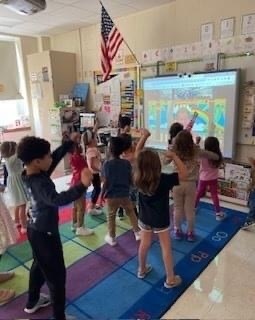 students standing on carpet in classroom watching SMARTBoard with author Jack Hartman