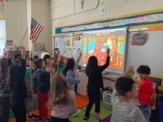 students standing on carpet in classroom watching SMARTBoard with author Jack Hartman